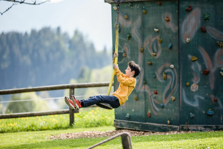 Ein Kind mit gelben T-Shirt sitzt auf einer Schaukel. Im Hintergrund ist eine Kletterwand und Wald zu erkennen.